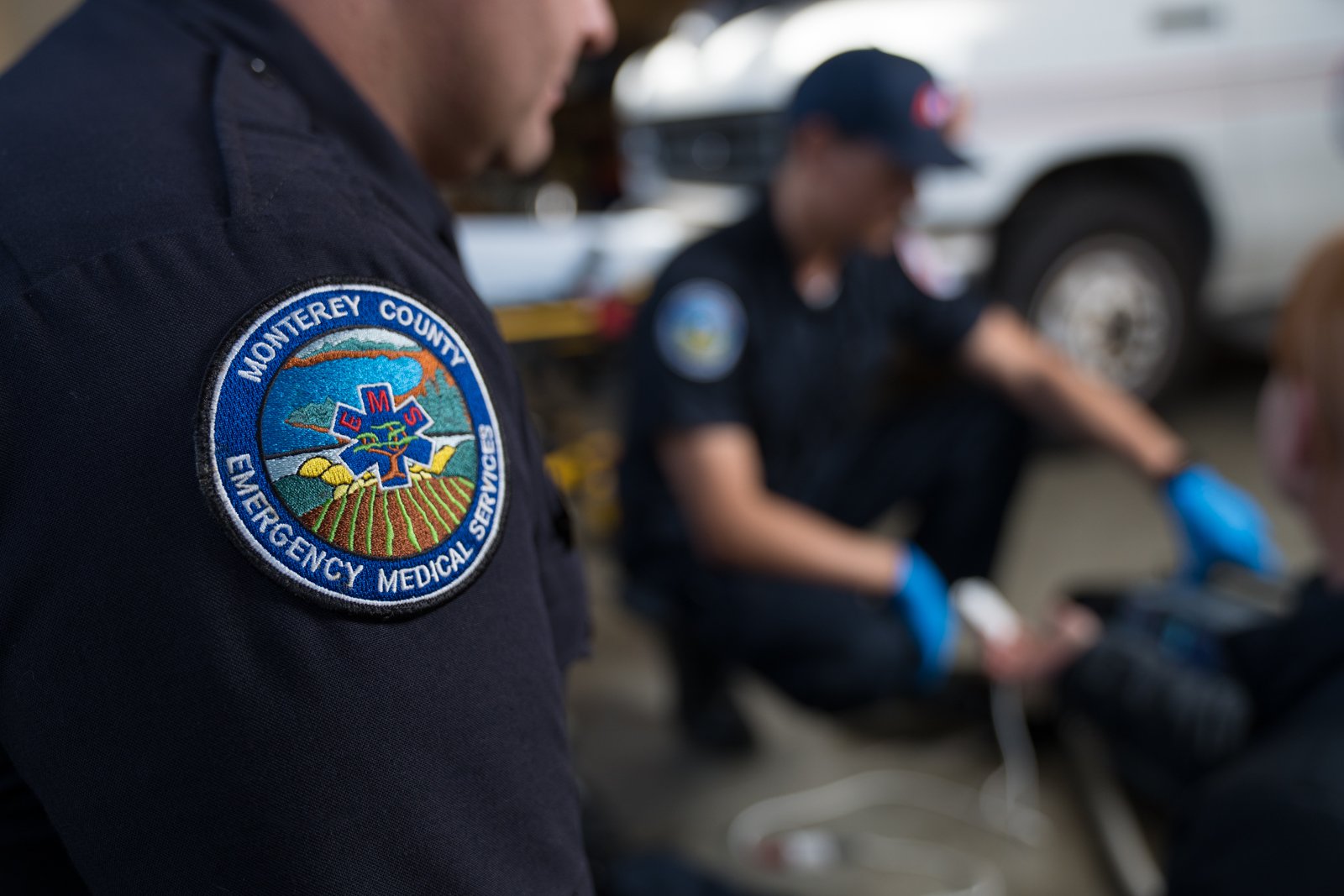 Close-up of Monterey County EMS badge on uniform, with paramedics assisting a patient in the background.