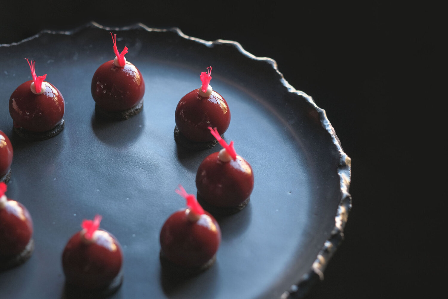 Artistic dessert presentation with glossy red spheres on a dark plate, each topped with a red garnish, against a dark background.