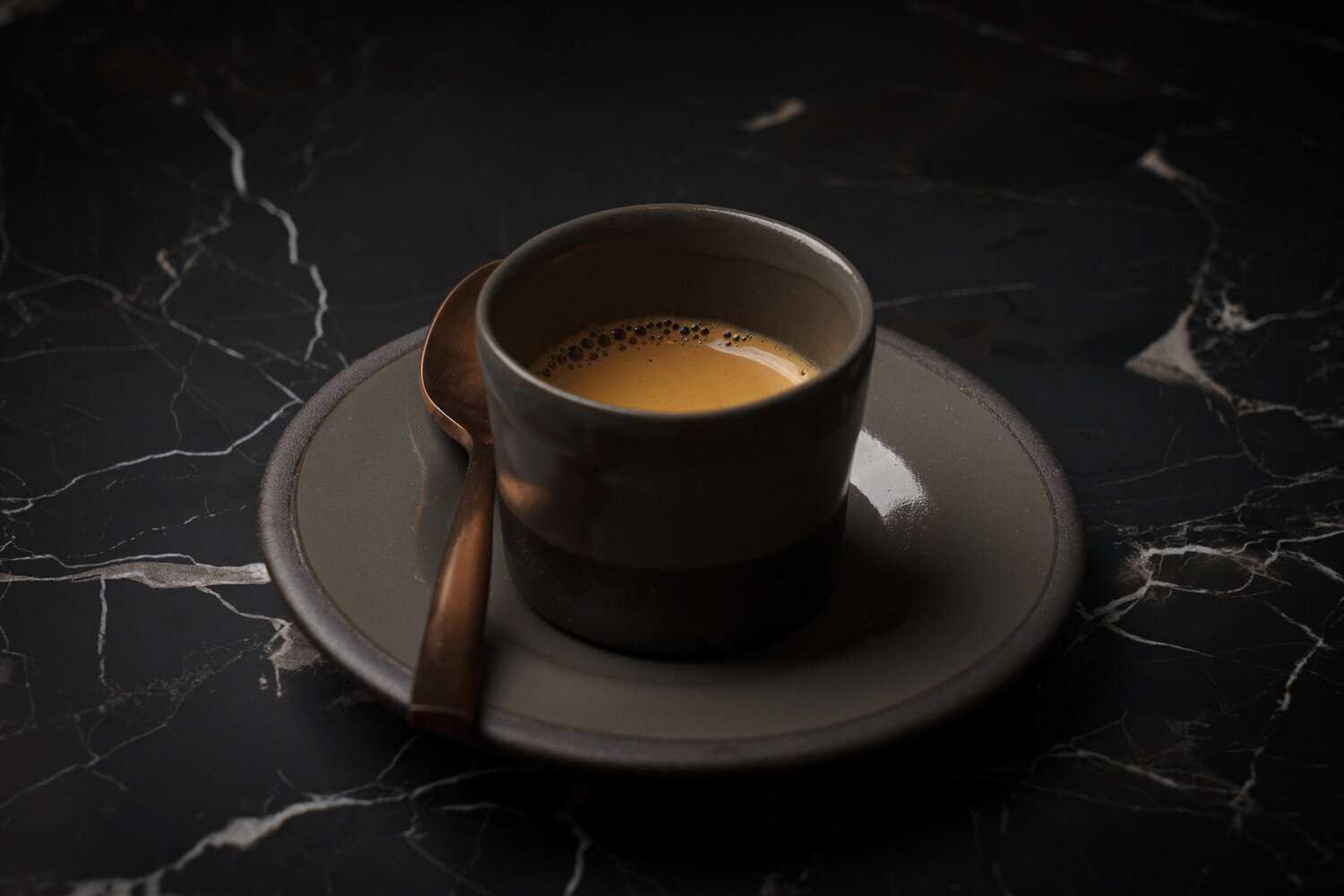 A cup of espresso on a saucer with a spoon, on a dark marble table.