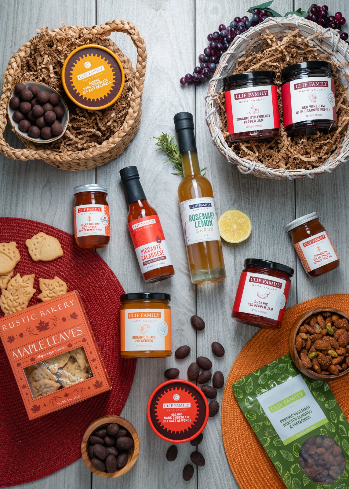 Assorted gourmet products including jams, sauces, almonds, and cookies displayed on a wooden surface with decorative baskets and mats.
