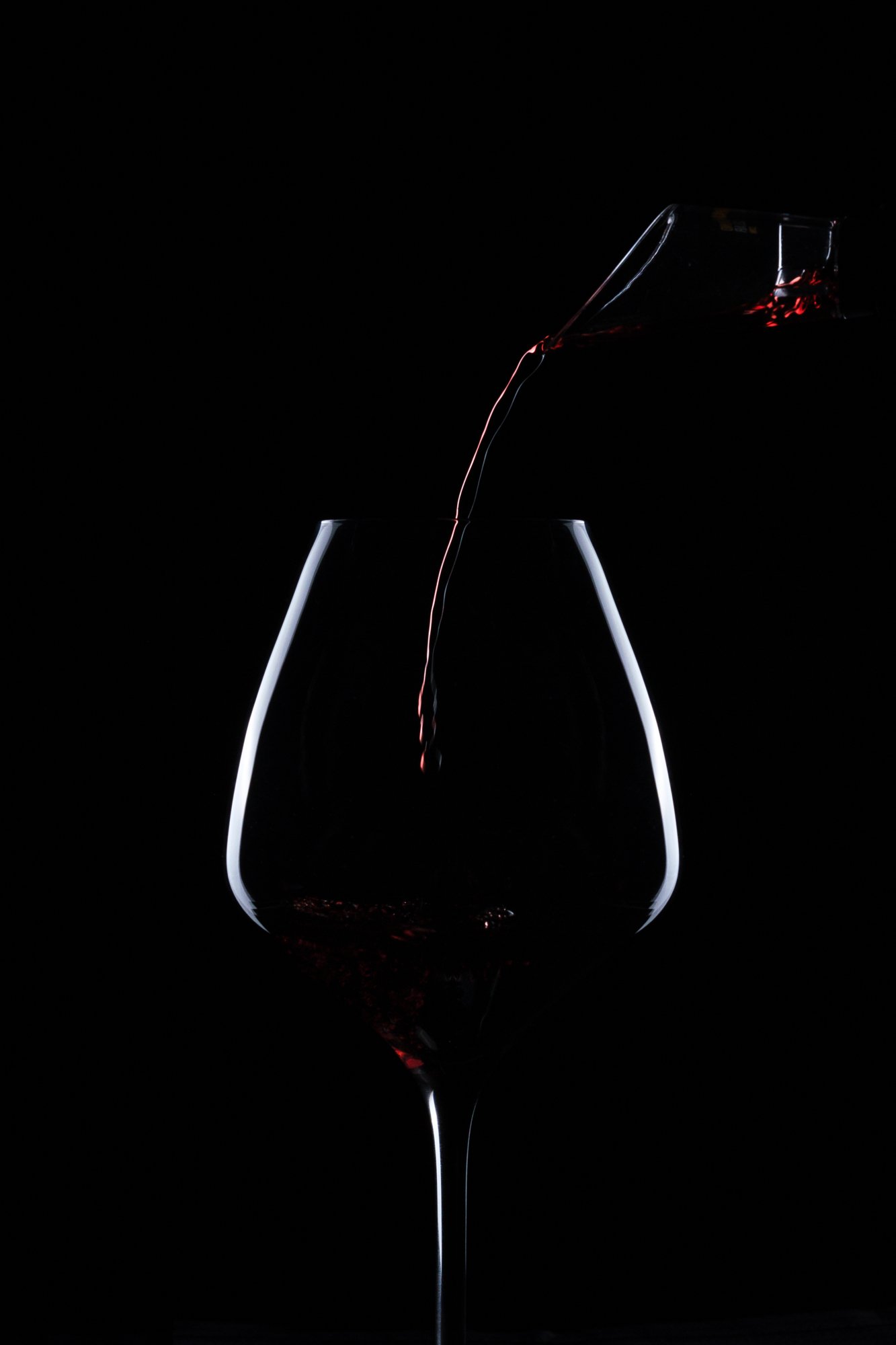 Dark silhouette of red wine being poured into a wine glass against a black background.