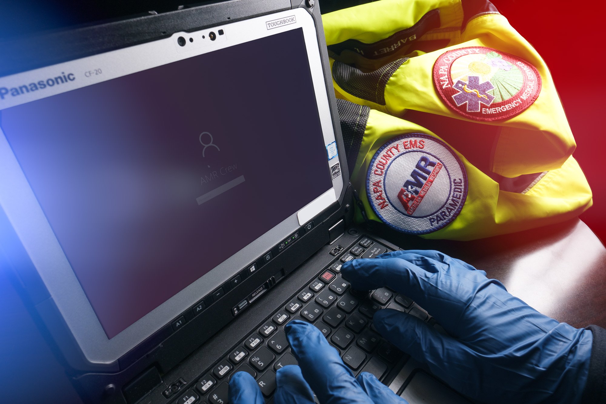 Person wearing blue gloves typing on a Panasonic Toughbook laptop, with emergency medical services (EMS) patches and a yellow safety jacket in the background.