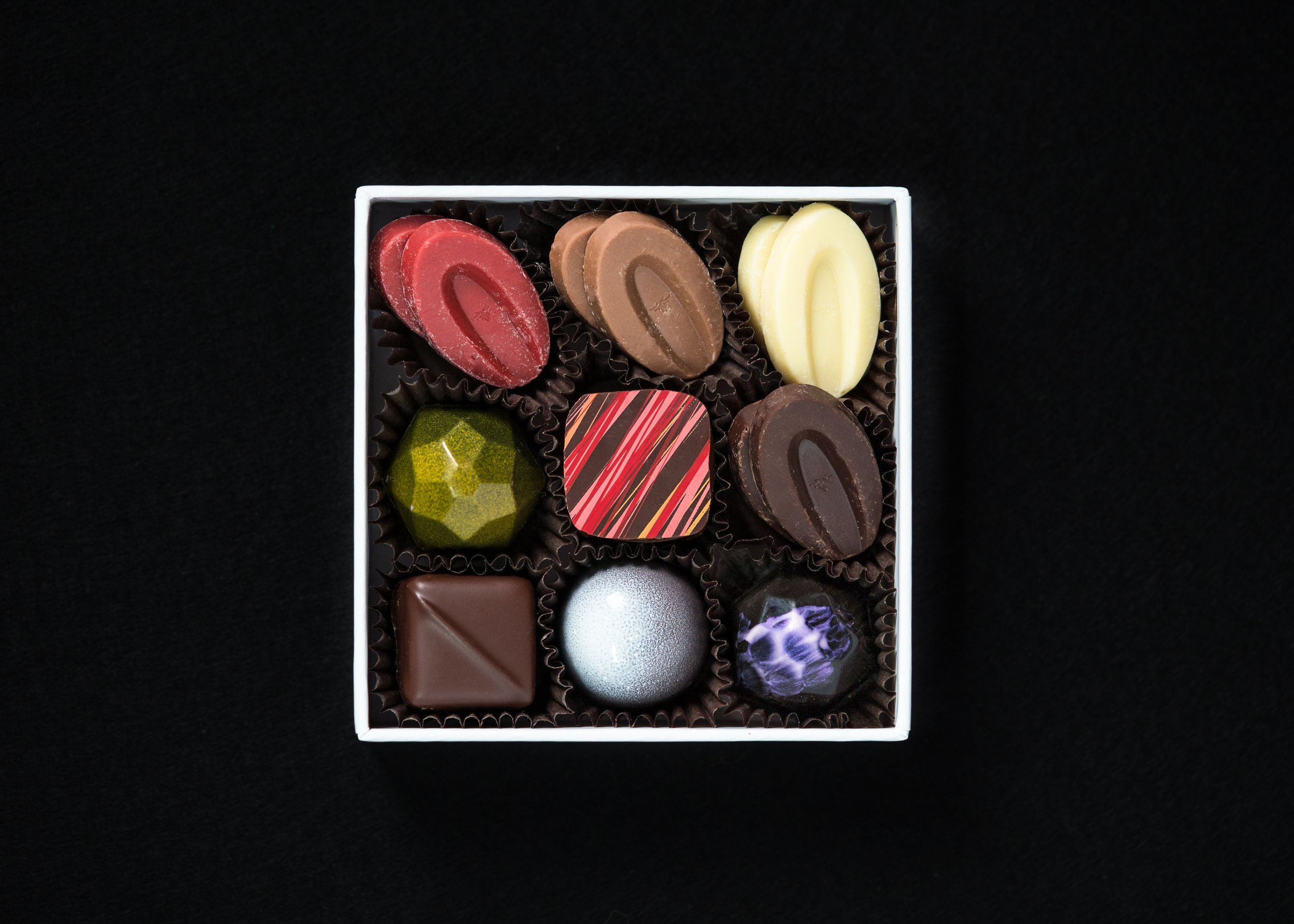 A box of assorted gourmet chocolates on a black background, featuring different shapes and colors such as red, brown, white, green, striped, and metallic designs.