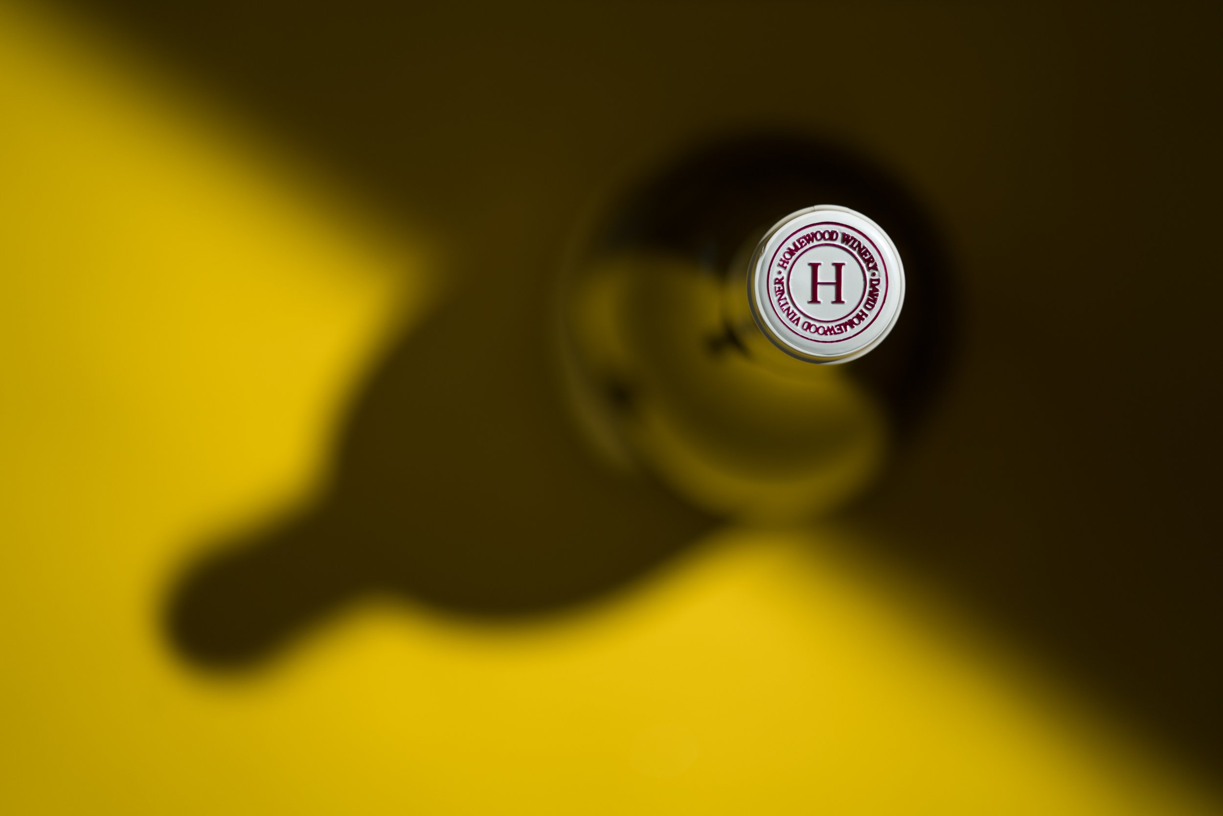 Overhead view of a bottle cap with the letter 'H' and surrounding text on a yellow background with shadows.