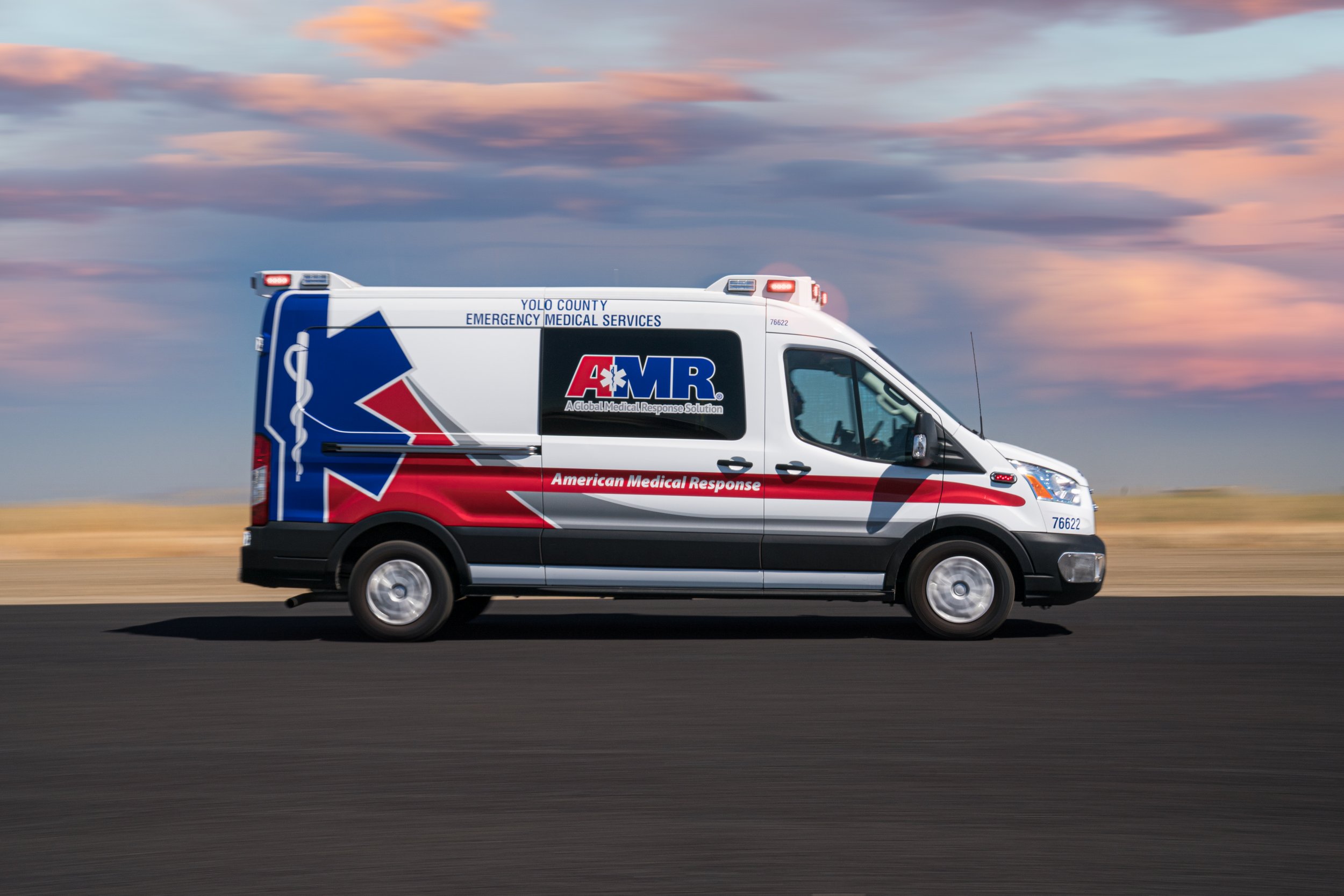 An American Medical Response (AMR) ambulance driving on a road with a sunset sky in the background.