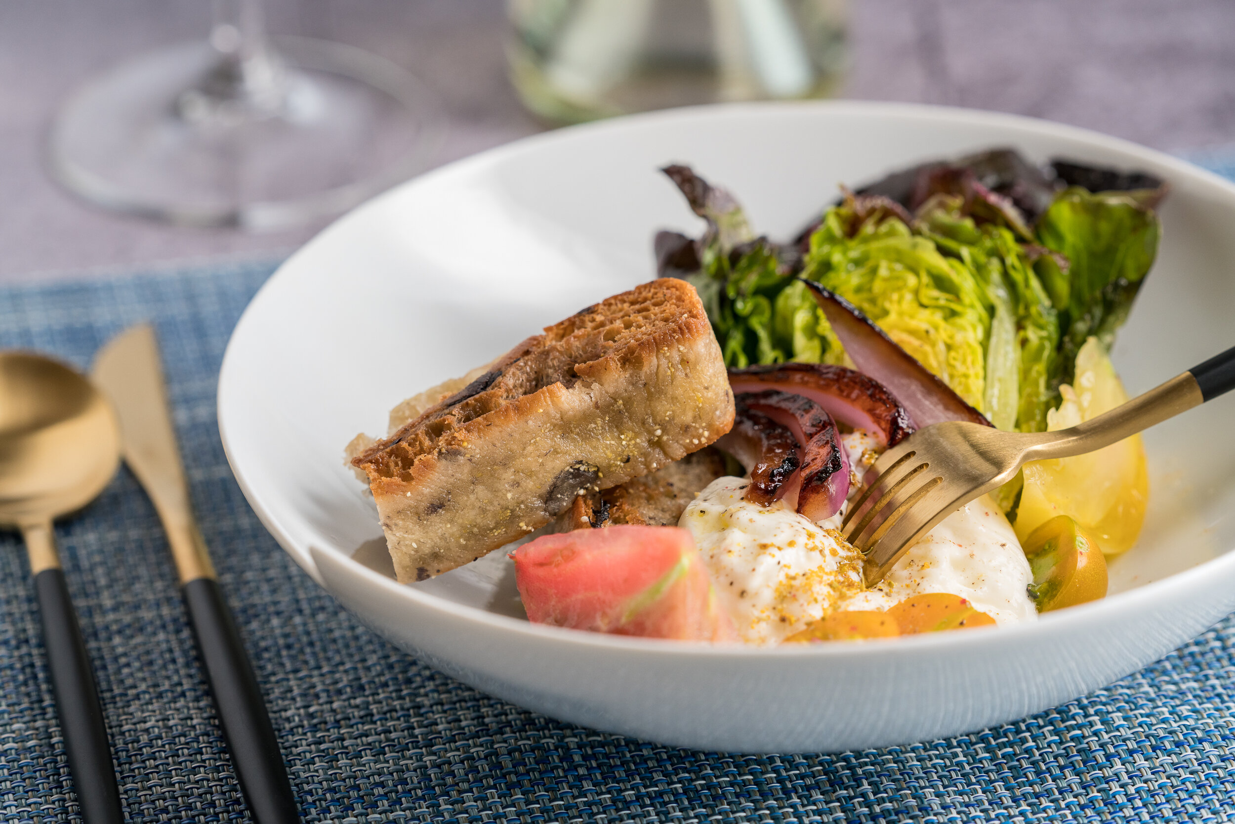 Gourmet salad with grilled bread, lettuce, red onion, heirloom tomatoes, and burrata cheese in a white bowl, placed on a blue textured placemat with gold utensils nearby.