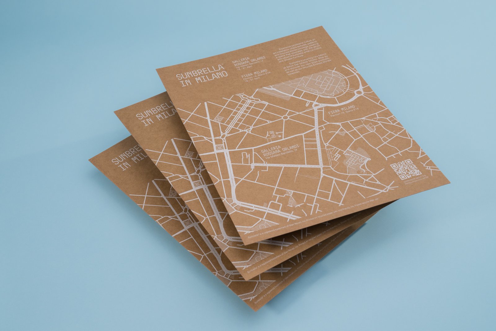 Stack of brown paper maps labeled 'Sunbrella in Milano' with white street map overlay on a blue background.