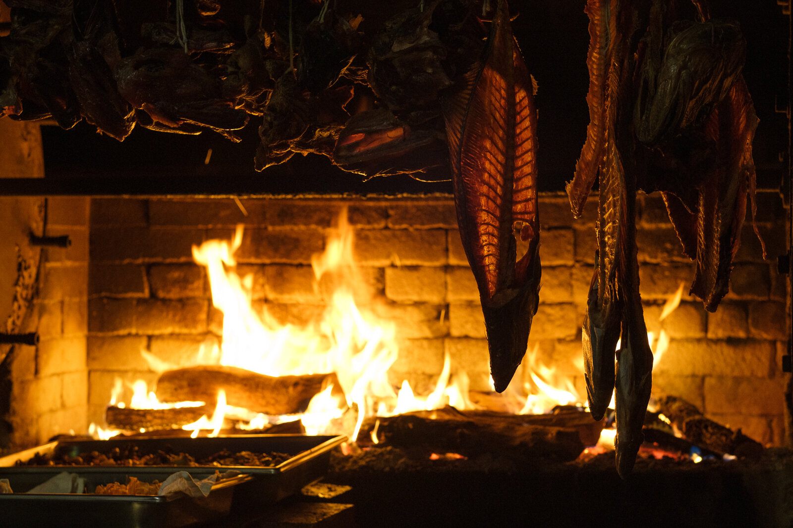 Fish smoking over an open wood fire in a brick fireplace with hanging racks.