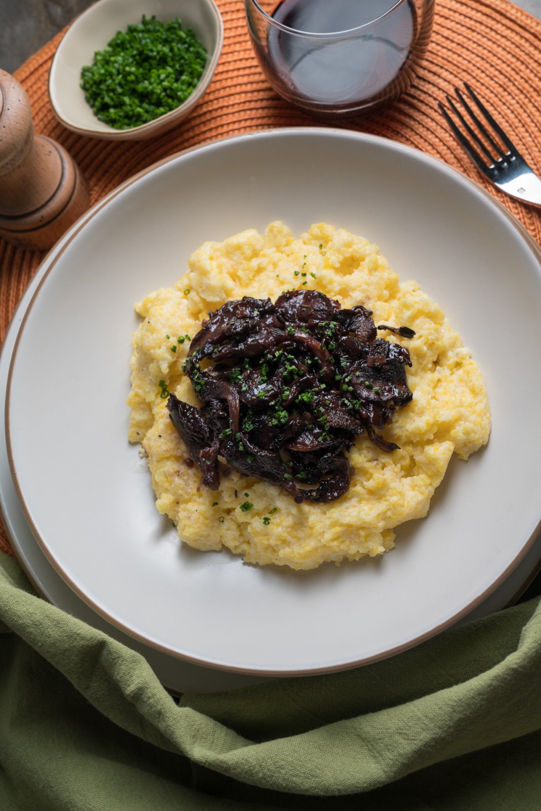 Plate of creamy polenta topped with sautéed mushrooms and chives, next to a glass of red wine, a small bowl of chopped chives, and a pepper grinder on an orange placemat.