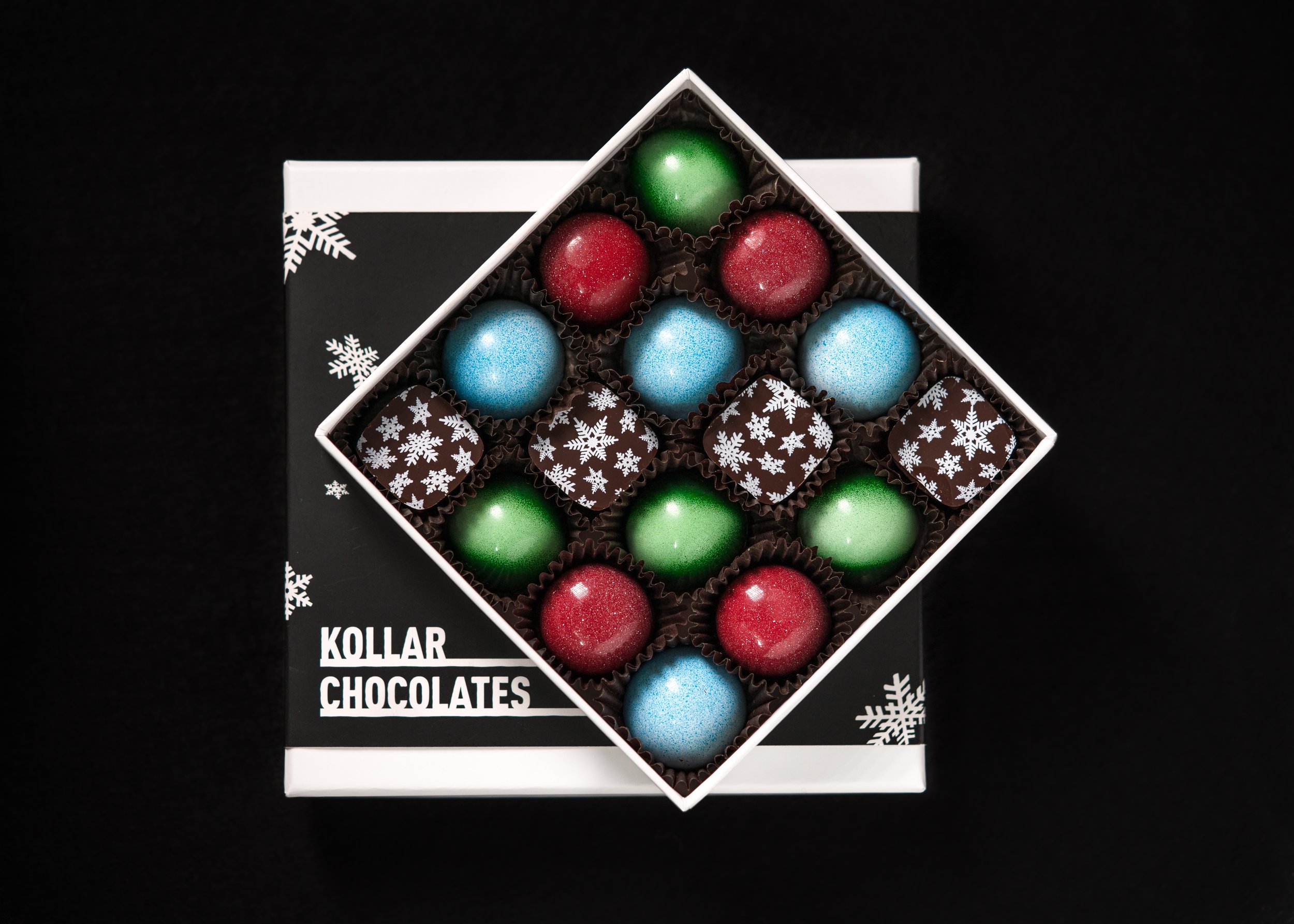 Box of assorted colorful chocolates with red, green, blue spheres and snowflake-patterned squares on a black background.
