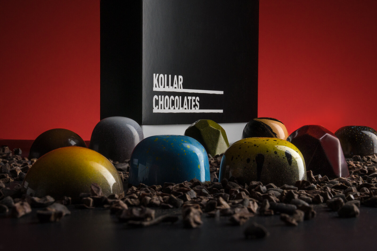 Gourmet chocolates with a glossy finish and various bright colors displayed in front of a black box labeled 'Kollar Chocolates', set against a red background, surrounded by cocoa nibs.