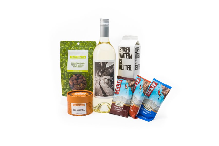 Assorted snack items including a bottle of white wine, boxed water, Clif bars, roasted nuts, and dark chocolate sea salt caramels.
