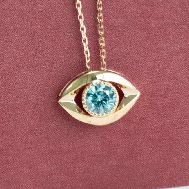 A striking memorial necklace with a big blue diamond set in a unique gold eye-shaped pendant, displayed on a mauve background.