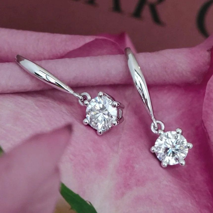 Elegant memorial diamond earrings with round-cut diamonds in a six-prong setting, set against a soft pink background.
