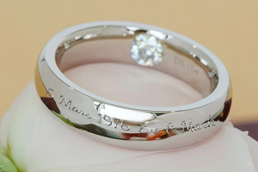 A modern minimalist white gold band with a brilliant diamond, engraved with "5 March 1976 Eva & Mark.