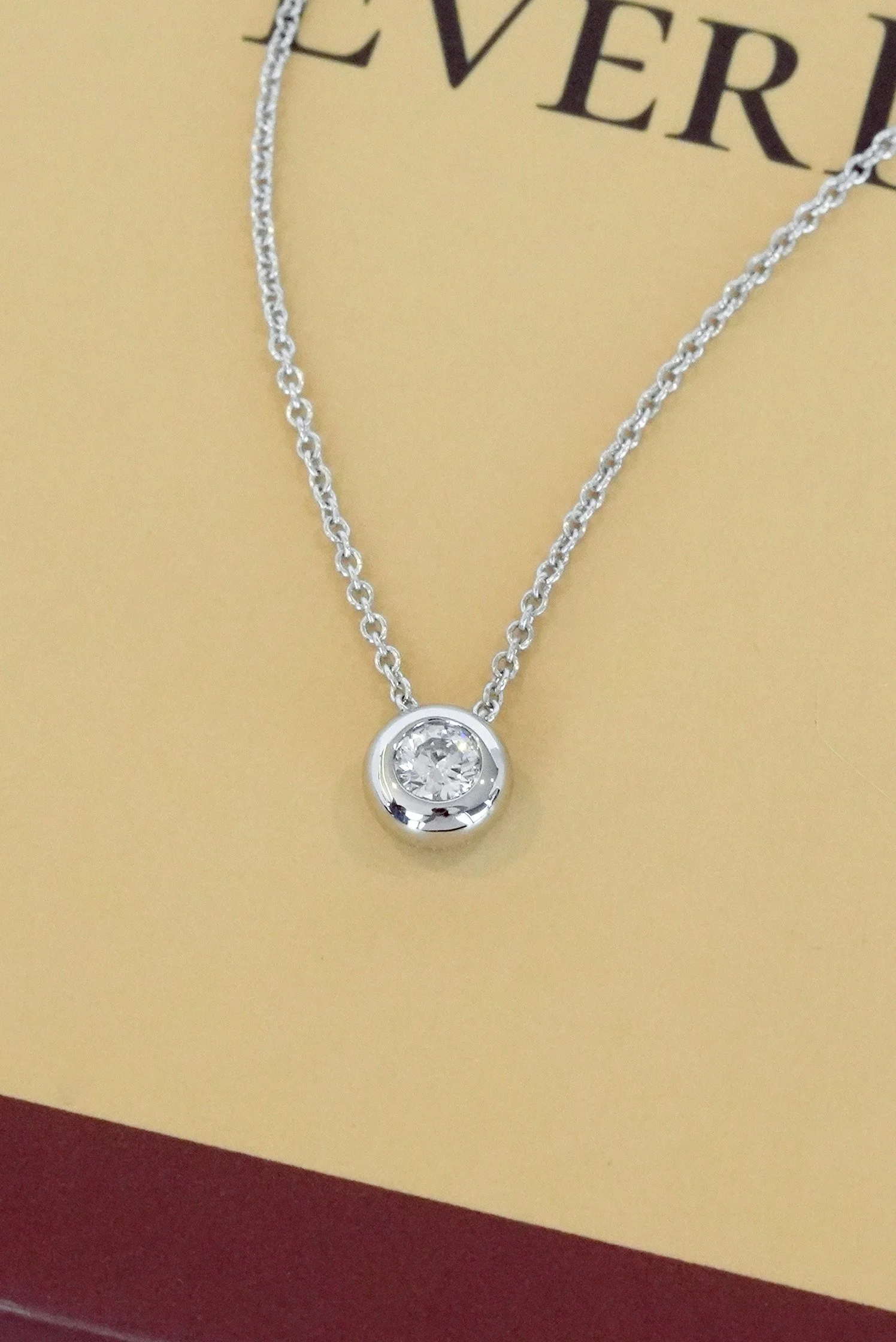 An 18k white gold necklace featuring a round diamond set in a smooth bezel, hanging gracefully on a delicate chain against a soft beige background.