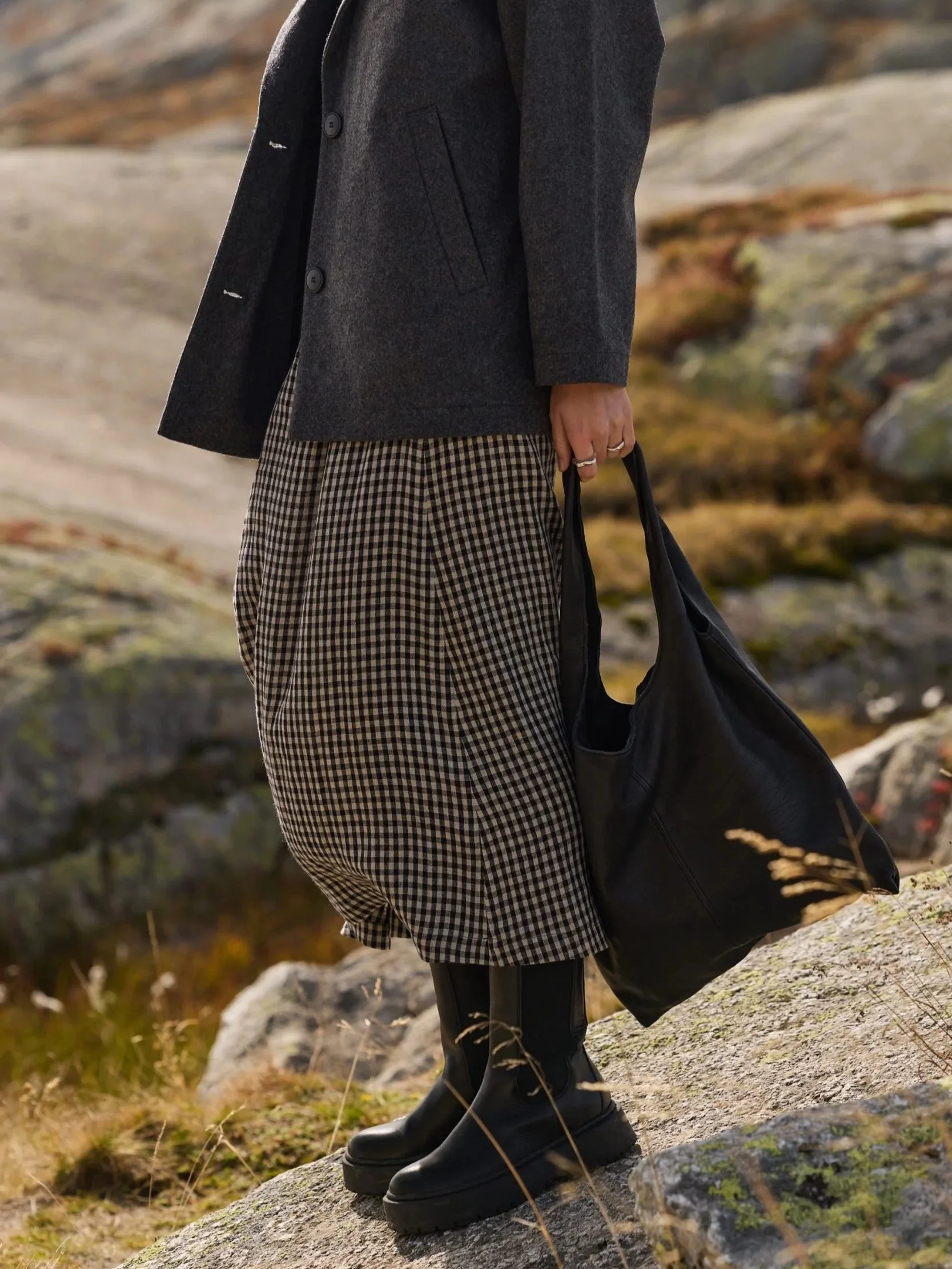 A simple tote made of vegetable-tanned goat leather in black, carried by hand with a plaid skirt and a gray wool coat in the Swiss Alps