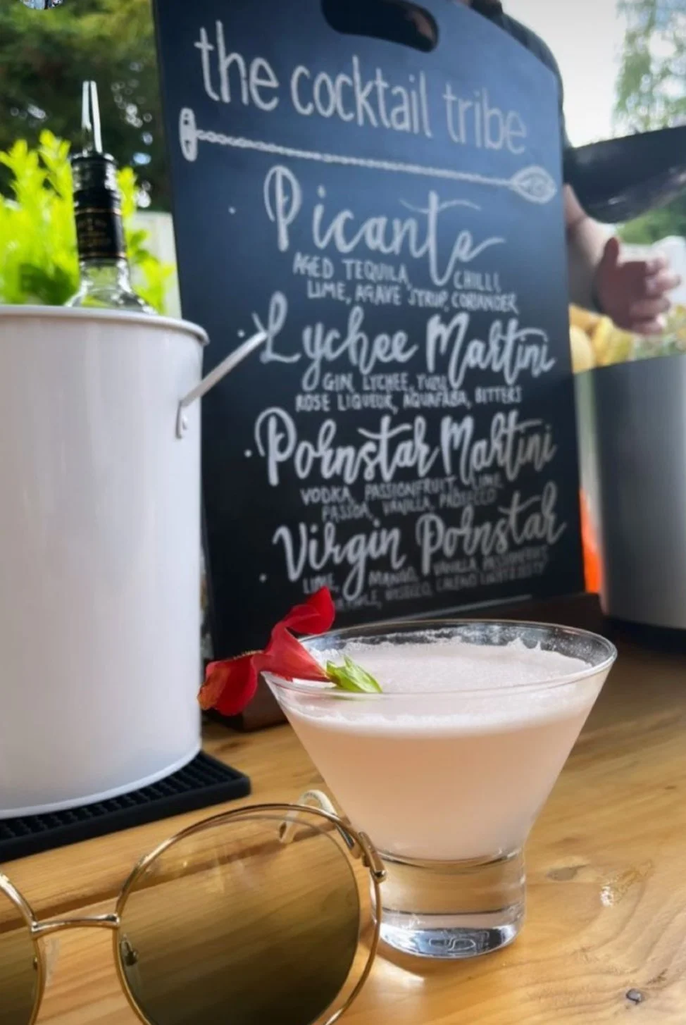 A lychee martini served in a martini glass garnished with an edible flower with a cocktail menu in the background and sunglasses in the foreground