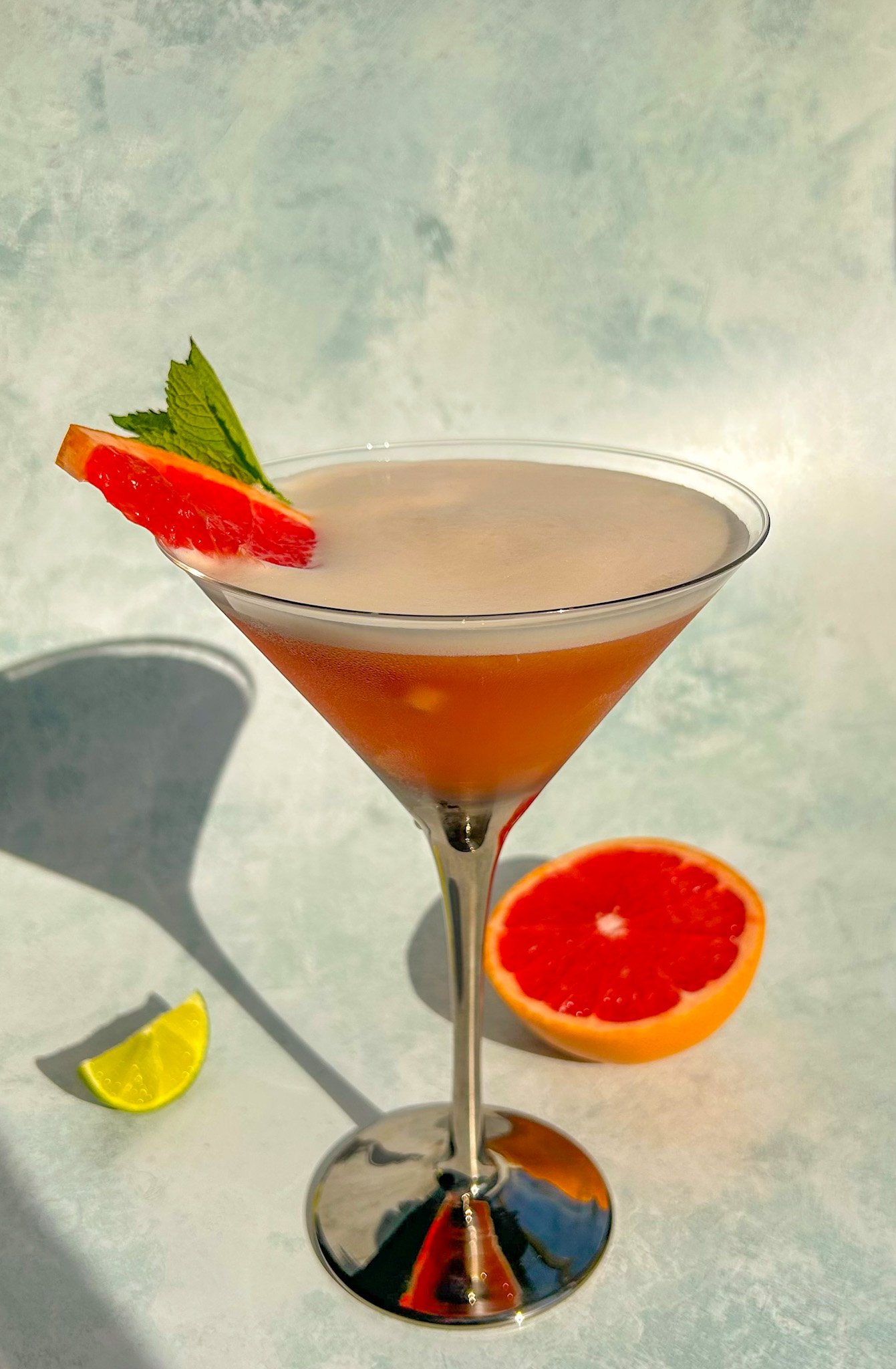A Grapefruit Mezcal Sour served in a martini glass and garnished with a grapefruit wedge and a mint leaf