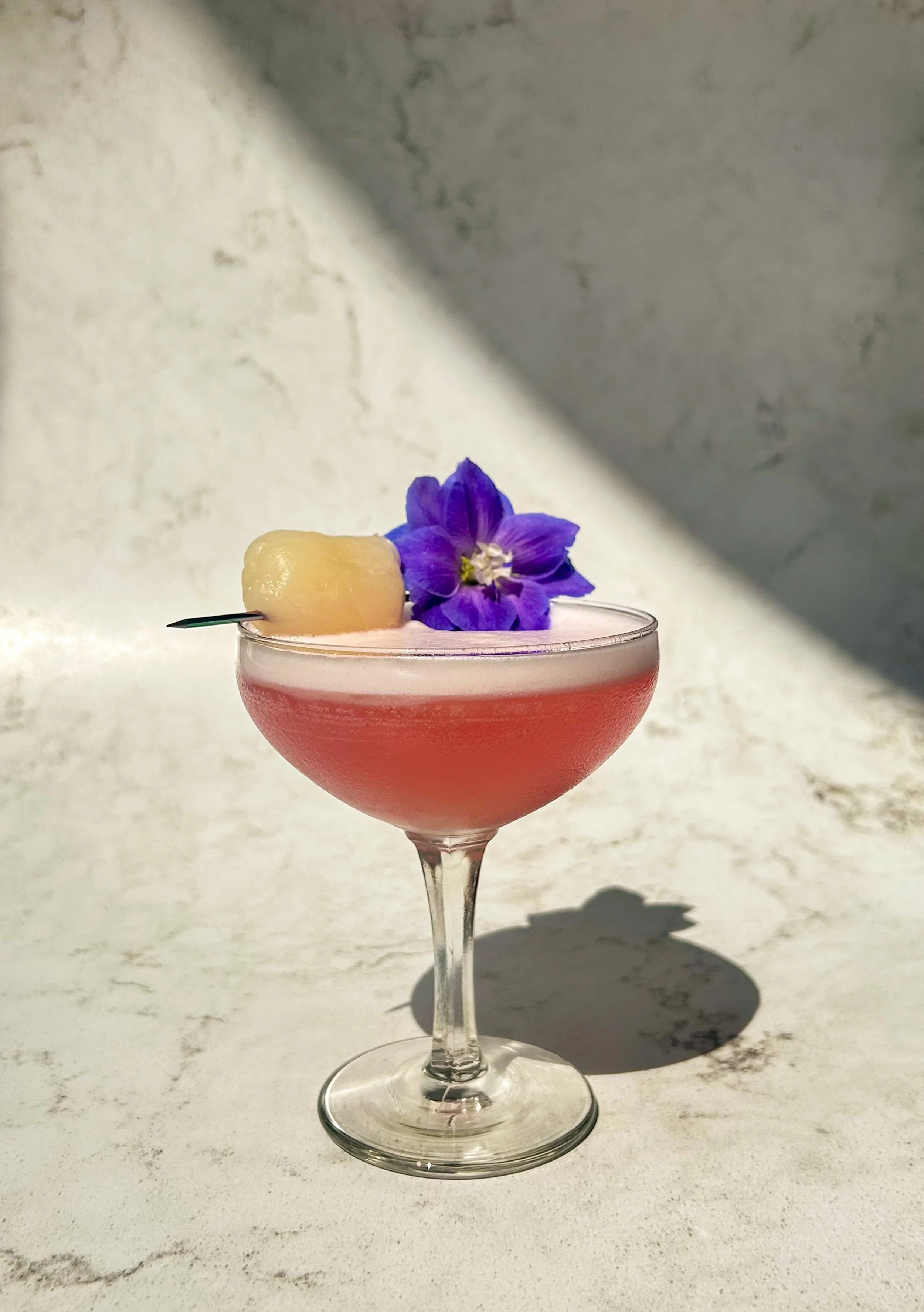 A lychee martini served in a coupe glass garnished with a fresh lychee and edible flower