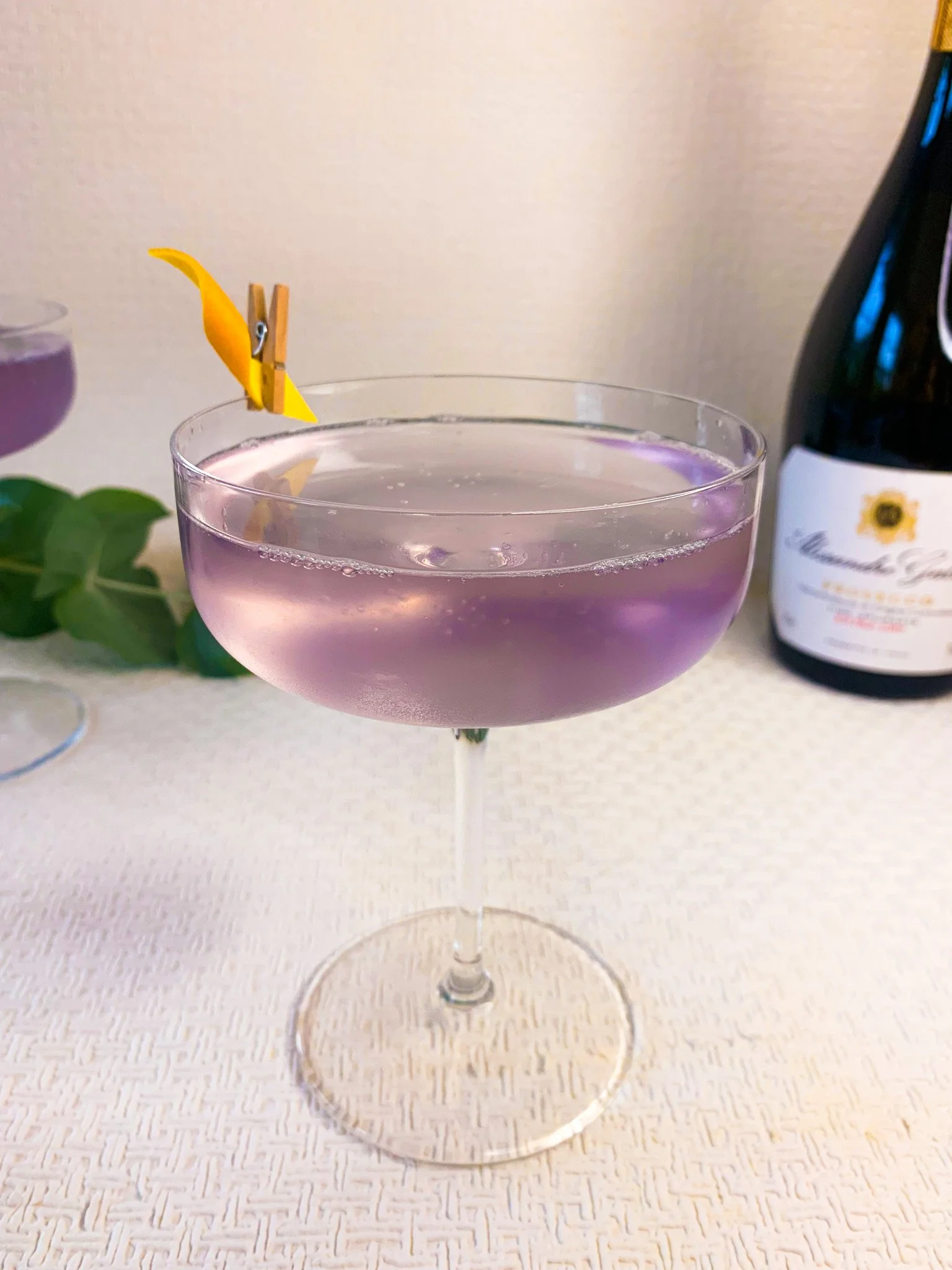 A lavender French 75 cocktail served in a coupe glass garnished with an orange rind