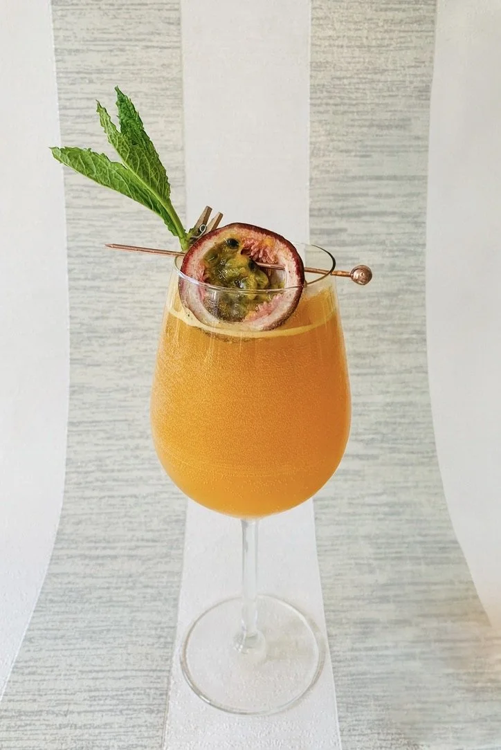 A Tropical Spritz cocktail served in a large wine gland and garnished with a skewered half passionfruit and a sprig of mint