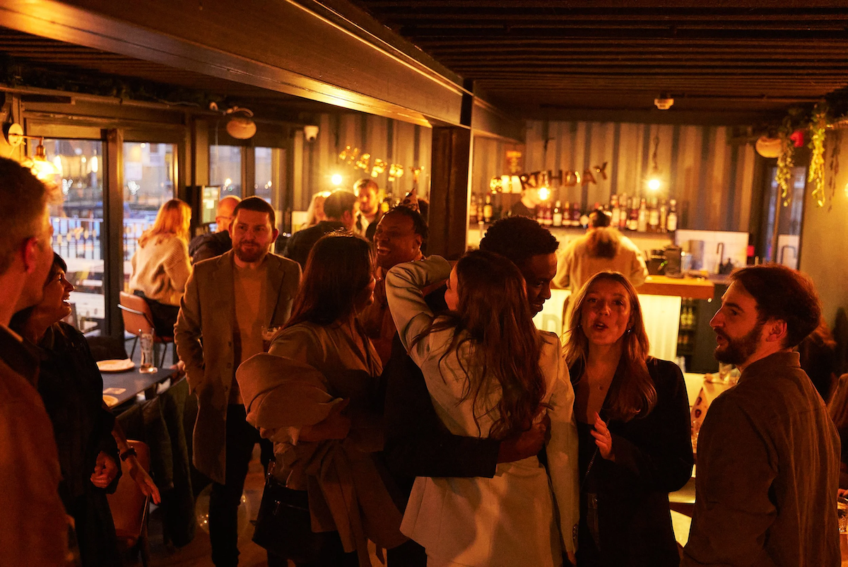 A group of people socializing and talking in a warmly lit bar or restaurant with a bar counter in the background.