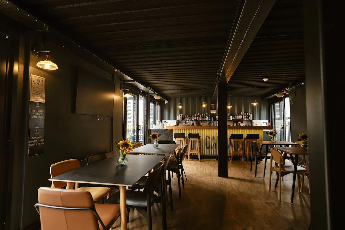 Interior of a modern bar and restaurant with wooden tables, chairs, and sunflower centerpieces, bar with bottles, black ceiling, and large windows.