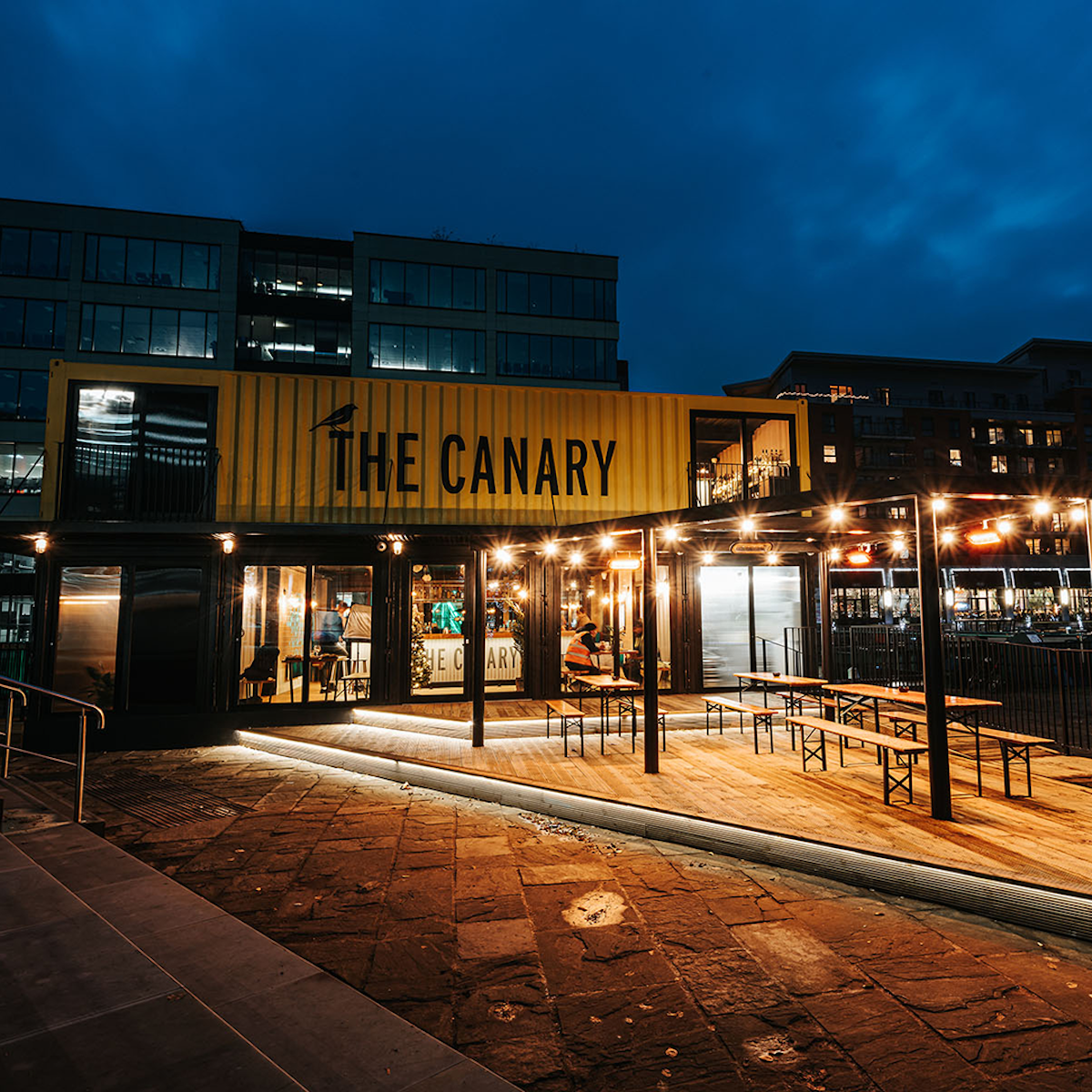 Night view of a modern building with a yellow shipping container sign reading 'The Canary' and outdoor seating area with tables and benches, illuminated by string lights.