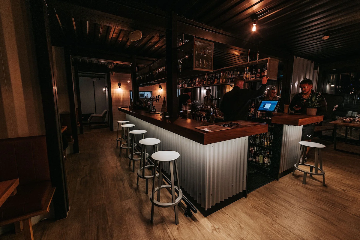 A dimly lit bar with a curved counter, bar stools, and shelves filled with bottles. Several people are behind the bar, and two are standing and talking. There are tables and chairs, and the floor is wooden.