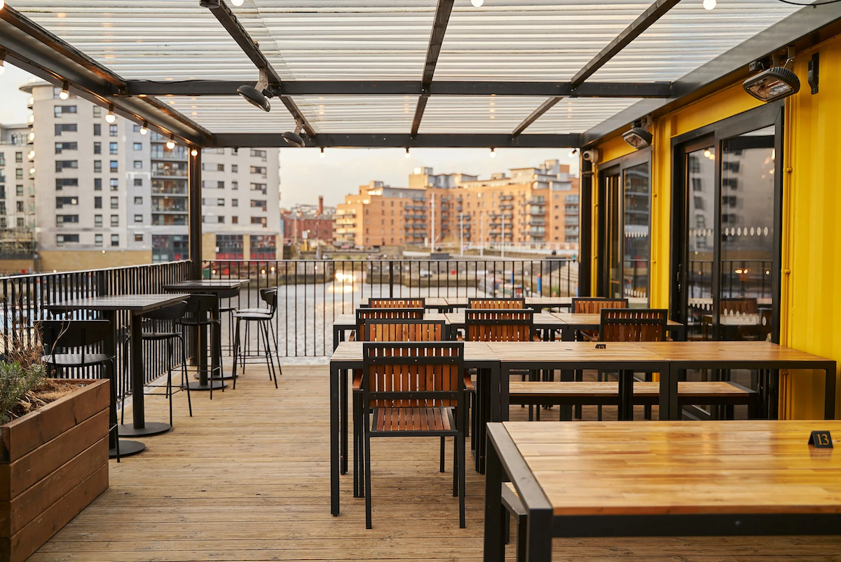 Empty outdoor patio with wooden tables and chairs, overlooking a city with tall residential buildings and a canal or river, during sunset.