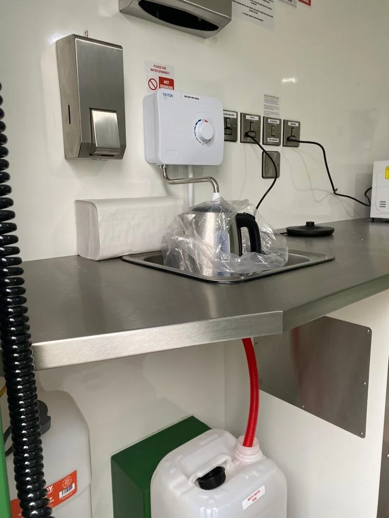 A laboratory or medical setting with a stainless steel counter, a white hot water dispenser, a whirlpool sterilizer, an electric kettle wrapped in plastic, electrical outlets with cords, a paper towel roll, and a water container with a red hose conne