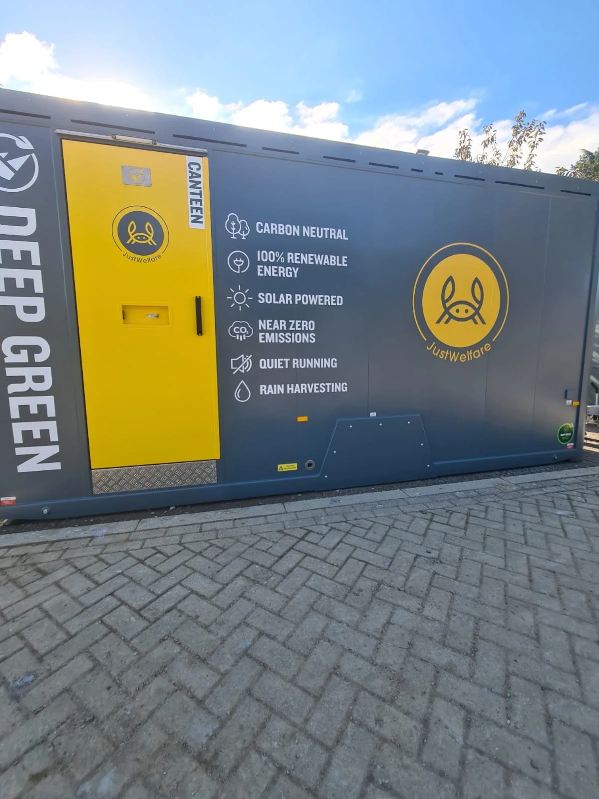 A large mobile recycling or waste container with information about its eco-friendly features, including being carbon neutral, renewable energy, solar powered, near zero emissions, quiet running, rain harvesting, and branding for JustWelfare. It is pl