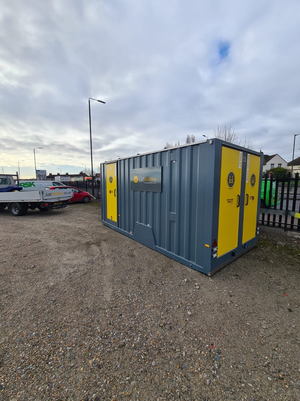 A blue and yellow mobile welfare unit in an outdoor parking area with gravel ground, surrounded by cars and fencing, under a cloudy sky.
