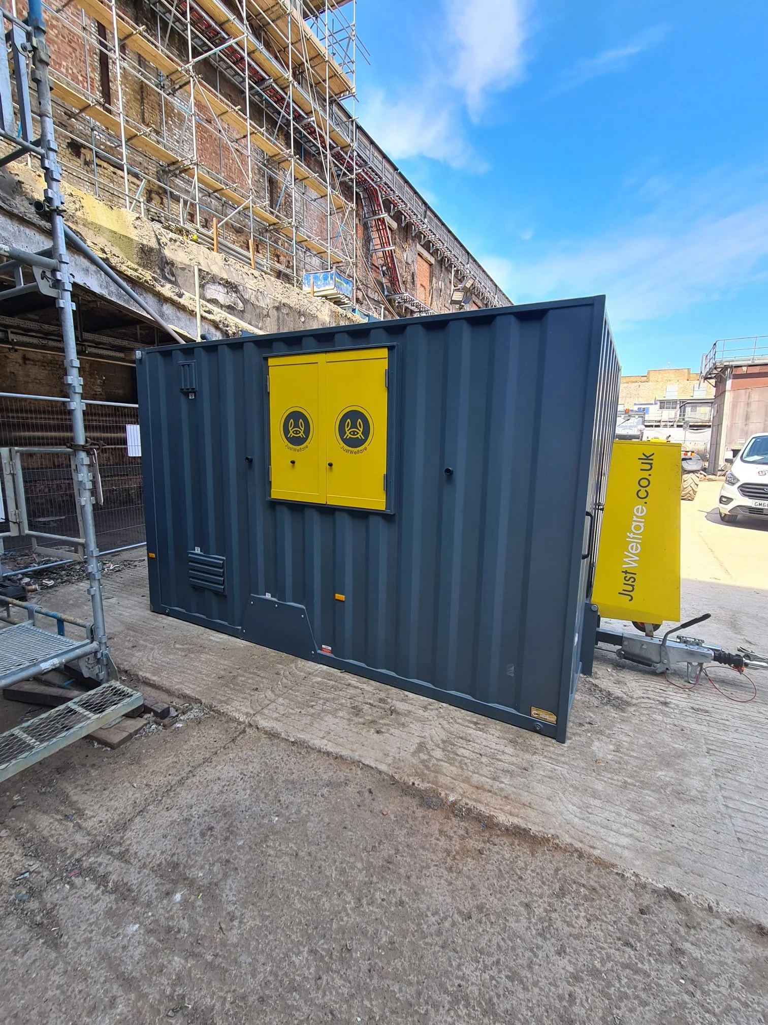 A blue shipping container with yellow access doors and warning signs, situated outdoors in a construction area with scaffolding, partially completed building, and parked cars in the background.