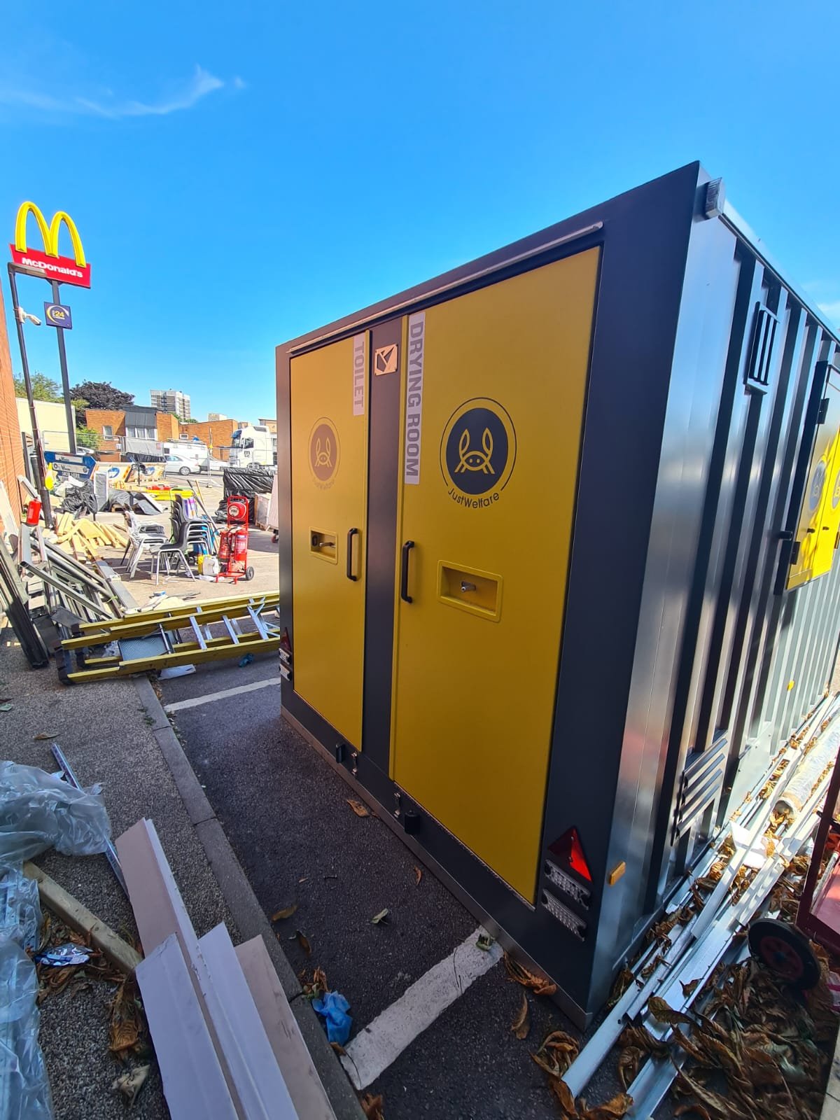 Yellow and gray utility cabinet labeled with signs for 'Drying Room' and 'Toilet' outside at a McDonald's parking lot with debris and equipment around.