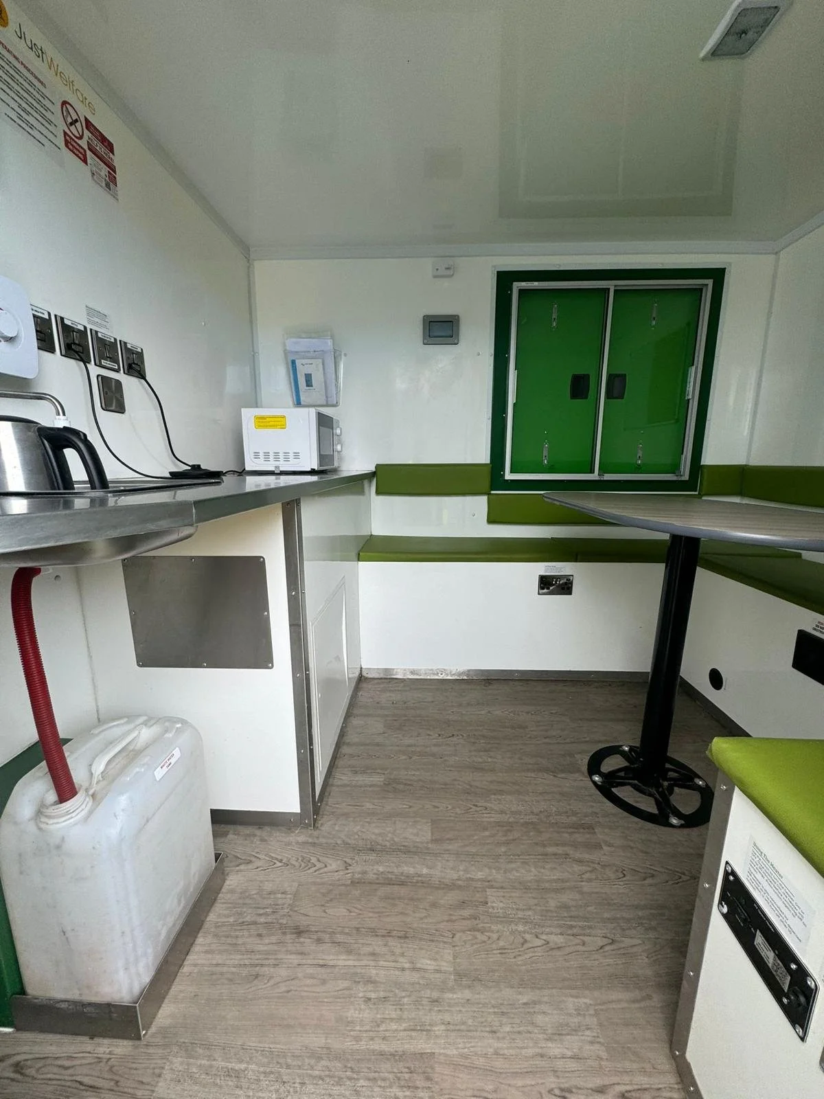 Interior of a small break room with a counter, microwave, kettle, and storage cabinets, featuring green trim and a wooden floor.