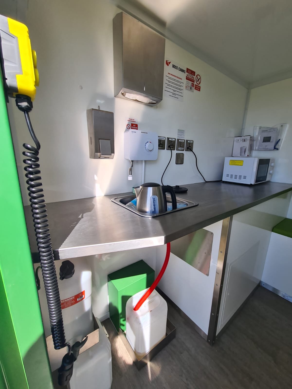 Small kitchenette area with microwave, kettle, and wall-mounted water heater, countertop with storage below, and wall-mounted electrical outlets, in a compact, industrial setting.