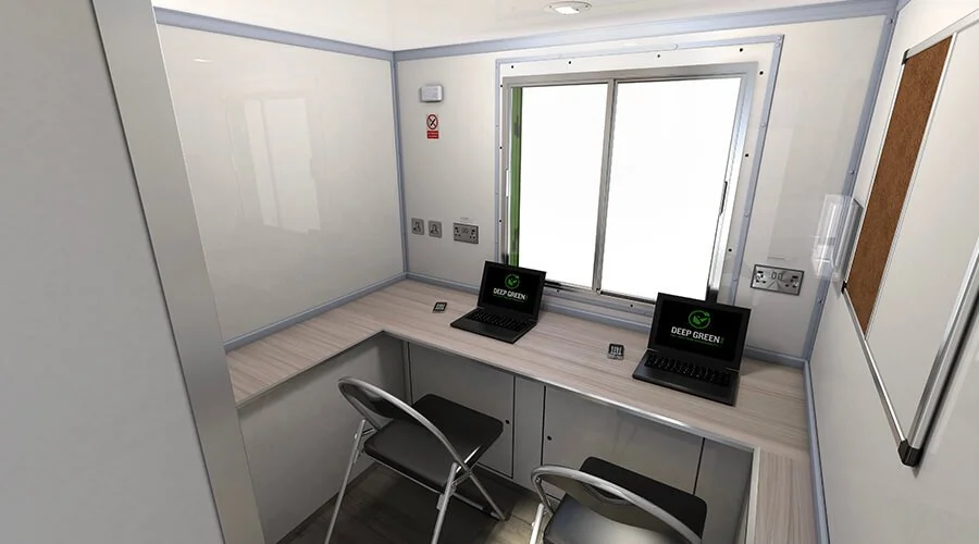 A small, clean office space with two laptops on a wooden desk, a window in the background, and two chairs in front of the desk. The laptops display a logo with the text 'DEEP GREEN.'