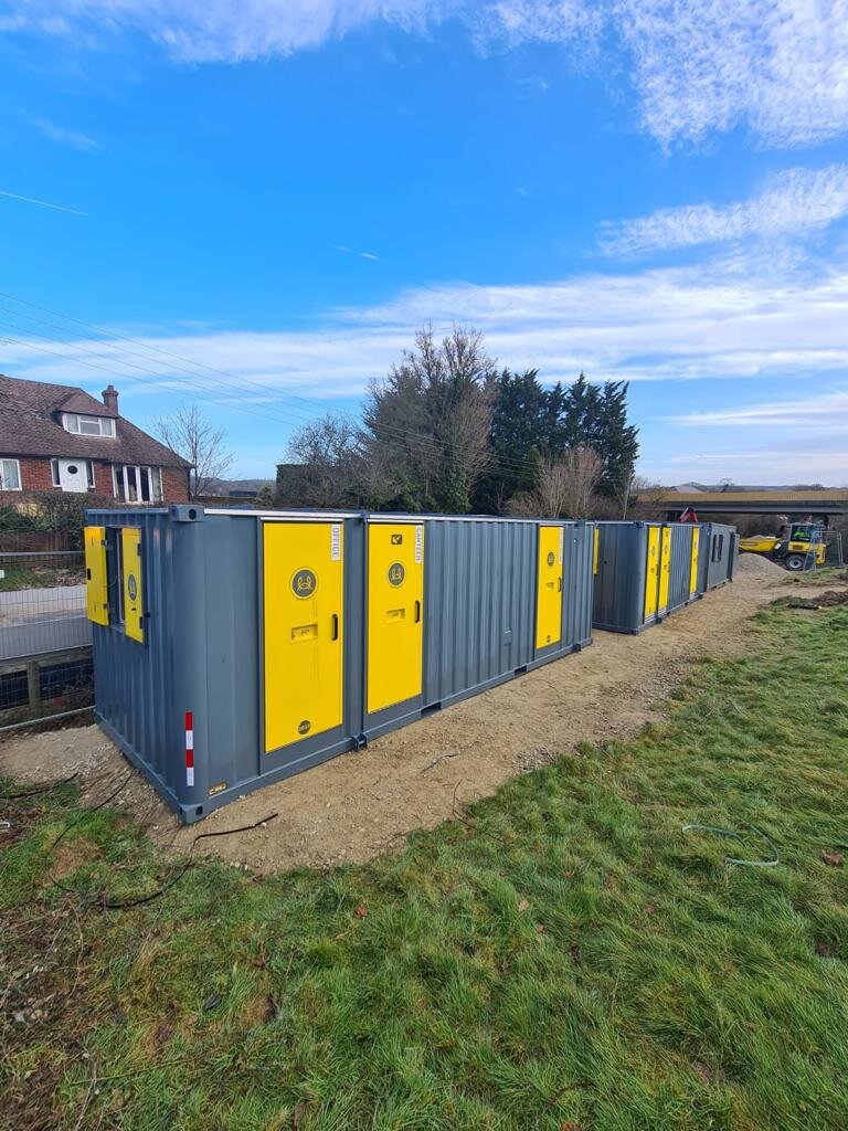 Multiple grey and yellow portable toilets or storage units lined up outdoors on a grassy and dirt area under a blue sky with clouds.