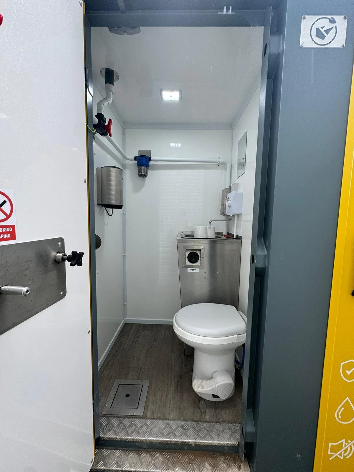Small industrial bathroom with toilet, stainless steel sink, water filtration system, and plumbing fixtures.