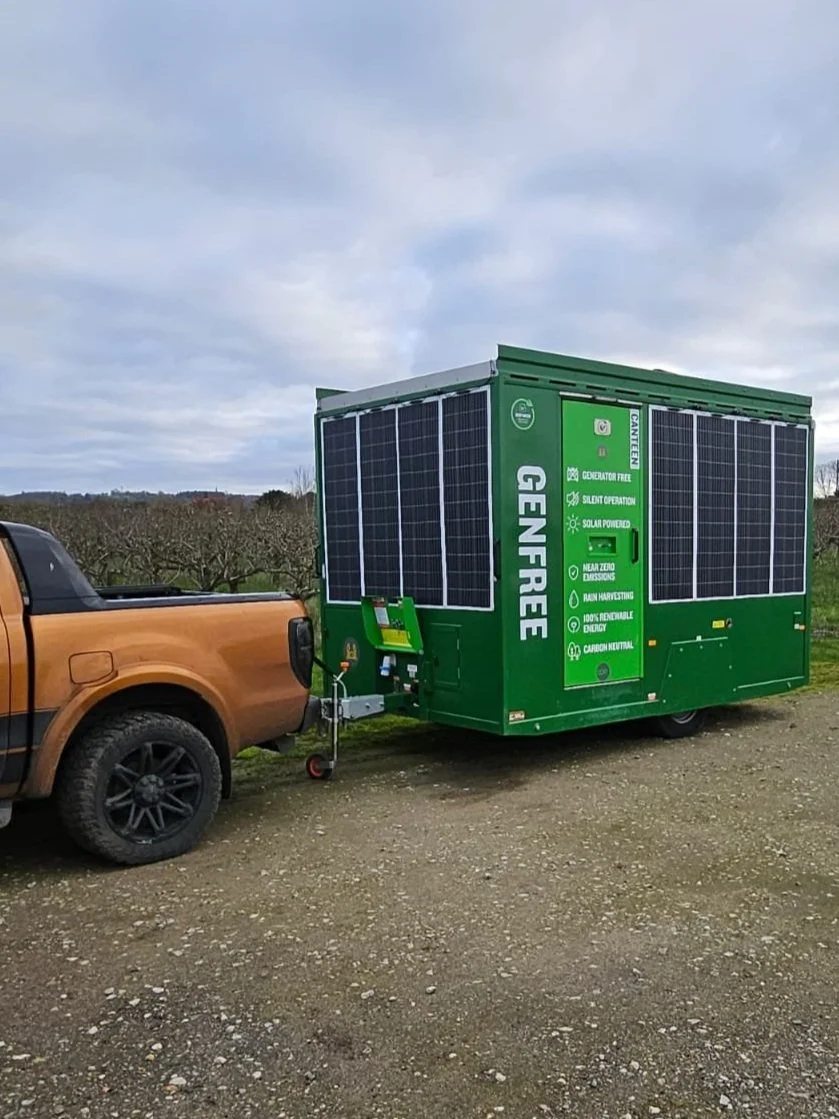 A truck pulling a green mobile generator with solar panels on top, in a rural area with trees and a cloudy sky in the background.