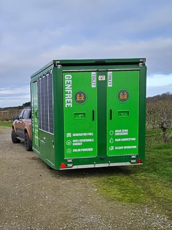 Green portable generator with solar panels, parked on gravel near fields, promoting renewable energy features.