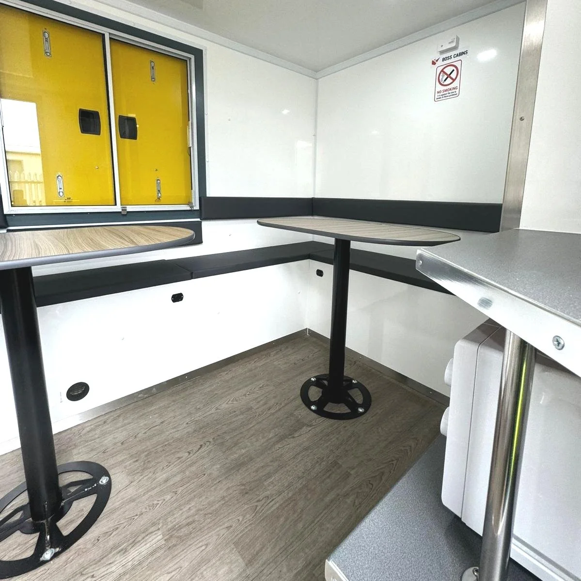 Interior of a small mobile food truck or trailer with two small tables and a bench seat, a yellow service window, and a no smoking sign on the wall.