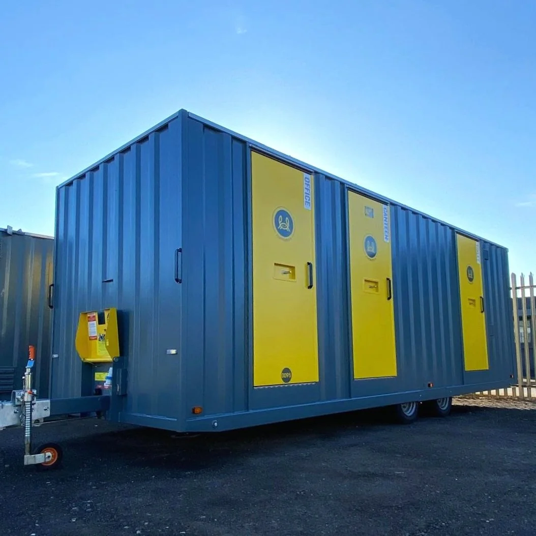 A large blue mobile generator building with yellow doors, situated outdoors on a paved surface, under a clear blue sky.