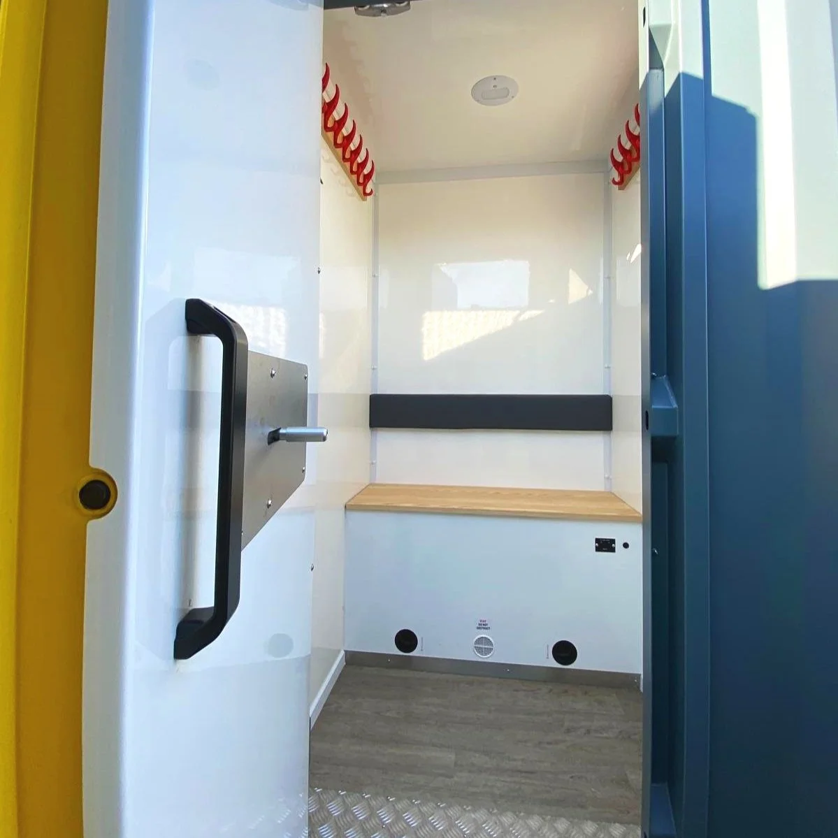 Inside a clean, empty mobile police or security cubicle with a black handle on the door, a wooden bench at the back, and red hook racks on the sides.