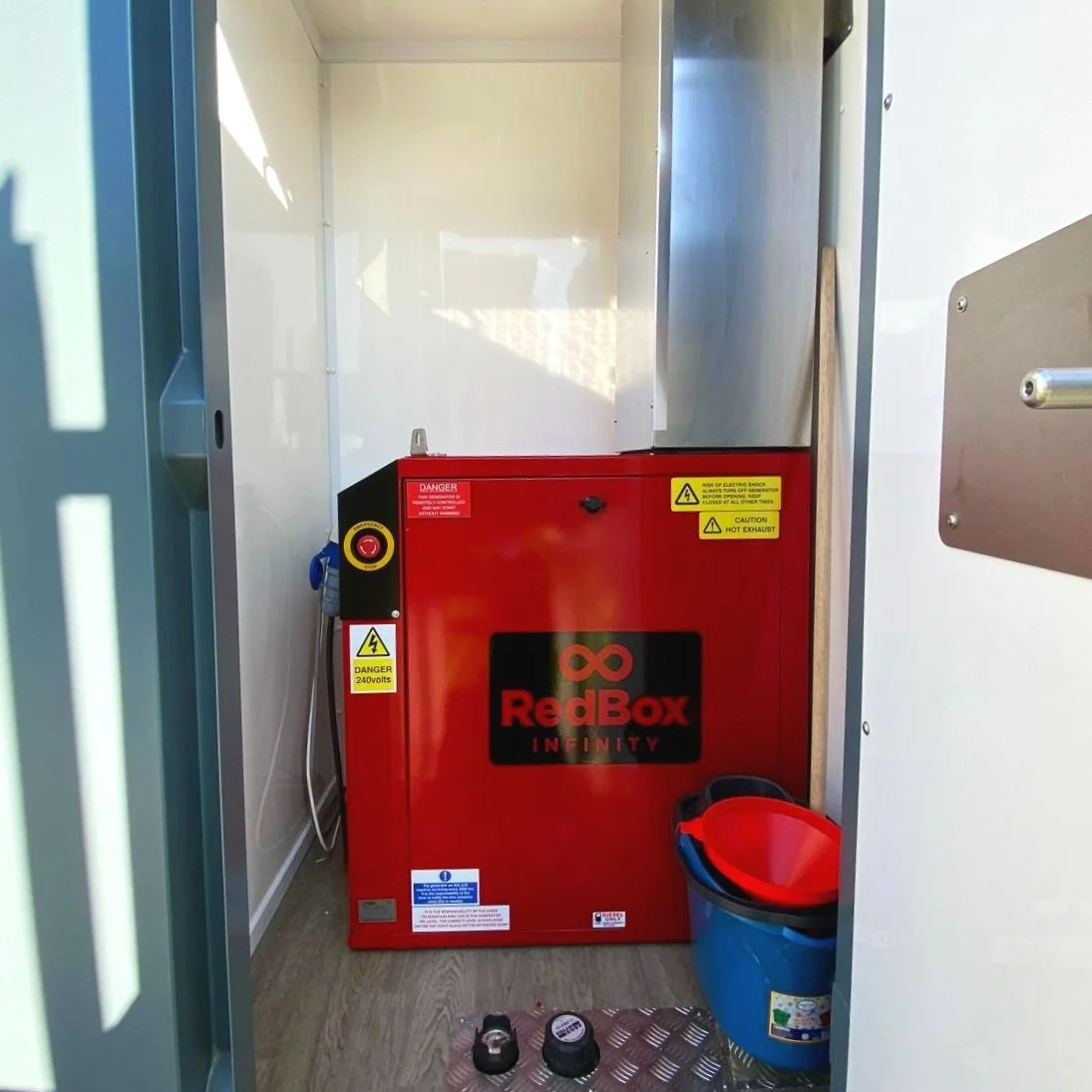 Electrical equipment in a small industrial or utility room, with a red box labeled 'RedBox Infinity' and safety warning labels, a bucket with a lid, and some small tools or components on the floor.