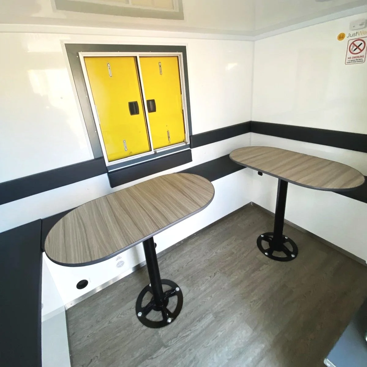 Two small tables with wooden tops and black metal bases in a corner of a room with white walls and a dark gray horizontal stripe. There is a yellow window on the wall and a no smoking sign in the upper right corner.