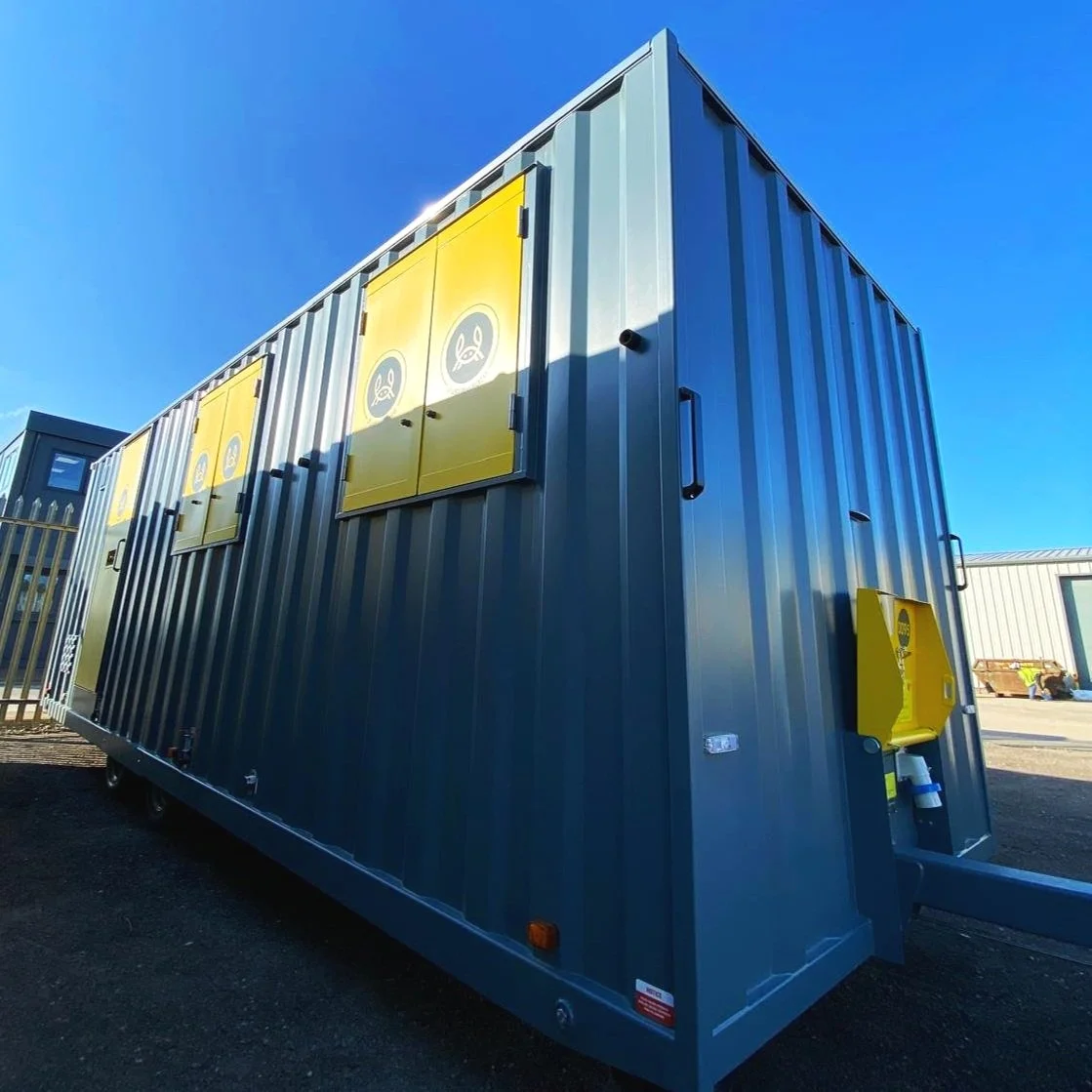 Large gray shipping container with yellow biohazard warning signs on the side and rear, located outdoors on a sunny day.