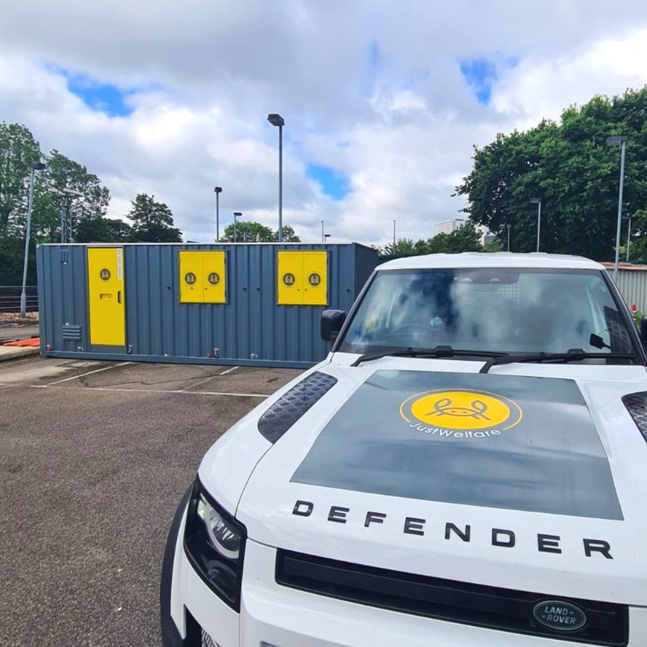 A Defendery vehicle with a logo on the hood, parked in a lot next to a blue utility container with yellow doors and panels under a cloudy sky with trees around.
